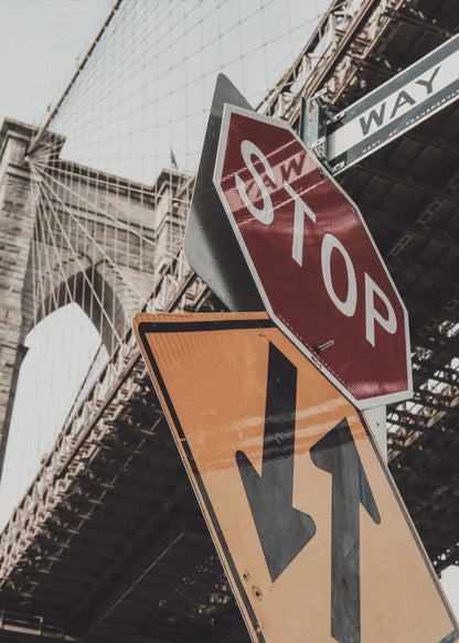 A low-angle photograph of the Brooklyn Bridge with a collection of street signs in the foreground. A red stop sign and a yellow sign with two arrows are prominent against the architectural details of the bridge. Print