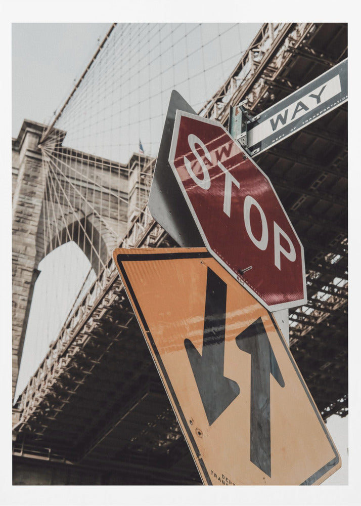 A low-angle photograph of the Brooklyn Bridge with a collection of street signs in the foreground. A red stop sign and a yellow sign with two arrows are prominent against the architectural details of the bridge. Print