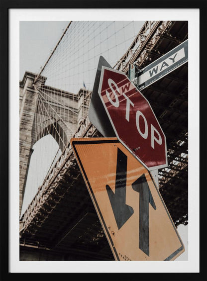 A low-angle photograph of the Brooklyn Bridge with a collection of street signs in the foreground. A red stop sign and a yellow sign with two arrows are prominent against the architectural details of the bridge. Print