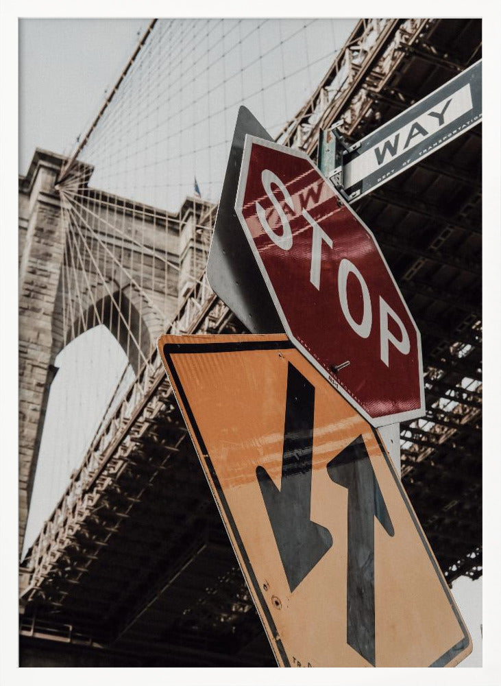 A low-angle photograph of the Brooklyn Bridge with a collection of street signs in the foreground. A red stop sign and a yellow sign with two arrows are prominent against the architectural details of the bridge. Print