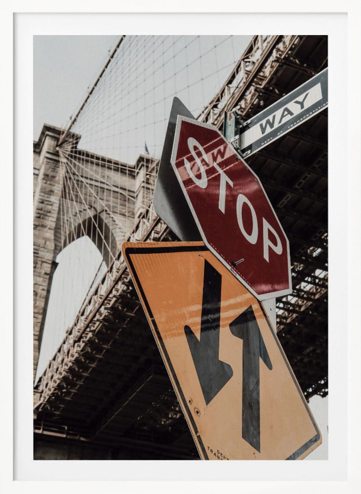 A low-angle photograph of the Brooklyn Bridge with a collection of street signs in the foreground. A red stop sign and a yellow sign with two arrows are prominent against the architectural details of the bridge. Print