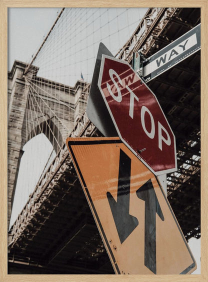 A low-angle photograph of the Brooklyn Bridge with a collection of street signs in the foreground. A red stop sign and a yellow sign with two arrows are prominent against the architectural details of the bridge. Print