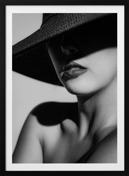 A dramatic black and white close-up photograph of a woman wearing a large, dark, wide-brimmed hat. The hat casts a deep shadow over the upper half of her face, obscuring her eyes and creating a sense of mystery. Only her nose, full lips, chin, neck, and shoulder are visible, highlighted by a strong light source. Decor