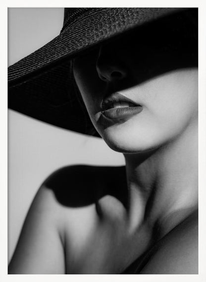 A dramatic black and white close-up photograph of a woman wearing a large, dark, wide-brimmed hat. The hat casts a deep shadow over the upper half of her face, obscuring her eyes and creating a sense of mystery. Only her nose, full lips, chin, neck, and shoulder are visible, highlighted by a strong light source. Decor