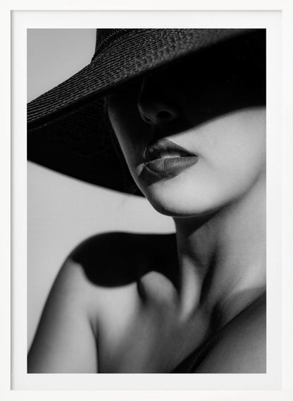 A dramatic black and white close-up photograph of a woman wearing a large, dark, wide-brimmed hat. The hat casts a deep shadow over the upper half of her face, obscuring her eyes and creating a sense of mystery. Only her nose, full lips, chin, neck, and shoulder are visible, highlighted by a strong light source. Decor