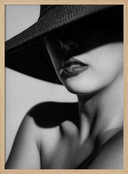 A dramatic black and white close-up photograph of a woman wearing a large, dark, wide-brimmed hat. The hat casts a deep shadow over the upper half of her face, obscuring her eyes and creating a sense of mystery. Only her nose, full lips, chin, neck, and shoulder are visible, highlighted by a strong light source. Decor