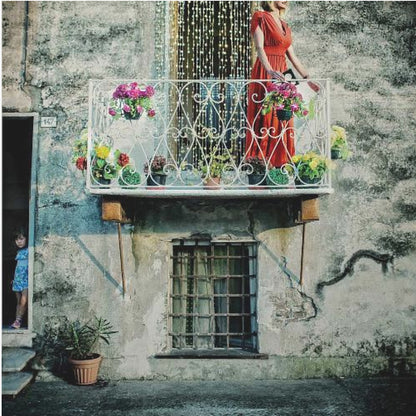 A woman in a vibrant red dress stands on an ornate white balcony filled with colorful potted flowers. The building has a weathered, gray stucco facade. To the left, a small child in a blue dress peeks out from a shadowed doorway, adding a layer of story to the rustic scene. Decor