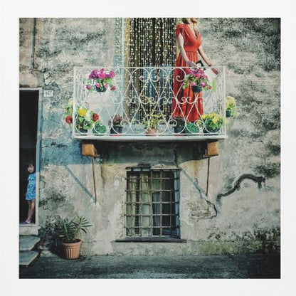 A woman in a vibrant red dress stands on an ornate white balcony filled with colorful potted flowers. The building has a weathered, gray stucco facade. To the left, a small child in a blue dress peeks out from a shadowed doorway, adding a layer of story to the rustic scene. Decor