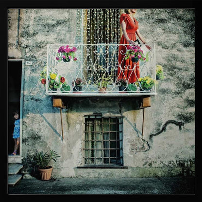 A woman in a vibrant red dress stands on an ornate white balcony filled with colorful potted flowers. The building has a weathered, gray stucco facade. To the left, a small child in a blue dress peeks out from a shadowed doorway, adding a layer of story to the rustic scene. Decor