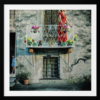 A woman in a vibrant red dress stands on an ornate white balcony filled with colorful potted flowers. The building has a weathered, gray stucco facade. To the left, a small child in a blue dress peeks out from a shadowed doorway, adding a layer of story to the rustic scene. Decor