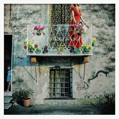 A woman in a vibrant red dress stands on an ornate white balcony filled with colorful potted flowers. The building has a weathered, gray stucco facade. To the left, a small child in a blue dress peeks out from a shadowed doorway, adding a layer of story to the rustic scene. Decor