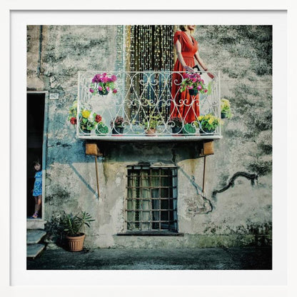 A woman in a vibrant red dress stands on an ornate white balcony filled with colorful potted flowers. The building has a weathered, gray stucco facade. To the left, a small child in a blue dress peeks out from a shadowed doorway, adding a layer of story to the rustic scene. Decor