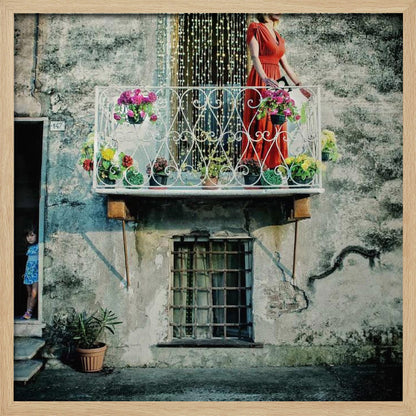 A woman in a vibrant red dress stands on an ornate white balcony filled with colorful potted flowers. The building has a weathered, gray stucco facade. To the left, a small child in a blue dress peeks out from a shadowed doorway, adding a layer of story to the rustic scene. Decor