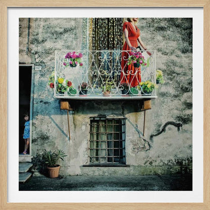 A woman in a vibrant red dress stands on an ornate white balcony filled with colorful potted flowers. The building has a weathered, gray stucco facade. To the left, a small child in a blue dress peeks out from a shadowed doorway, adding a layer of story to the rustic scene. Decor