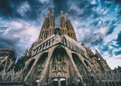 A dramatic, low-angle photograph of the Sagrada Familia in Barcelona, with its ornate stone facade and towering spires reaching up into a turbulent, dark blue and cloudy sky. The perspective emphasizes the grand scale and intricate detail of Gaudí's architecture. Print