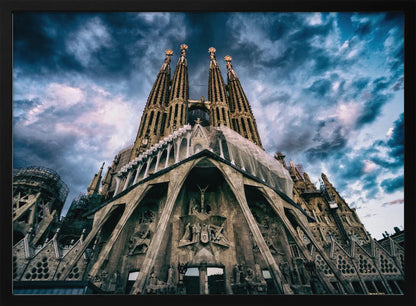 A dramatic, low-angle photograph of the Sagrada Familia in Barcelona, with its ornate stone facade and towering spires reaching up into a turbulent, dark blue and cloudy sky. The perspective emphasizes the grand scale and intricate detail of Gaudí's architecture. Print