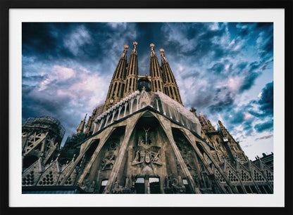 A dramatic, low-angle photograph of the Sagrada Familia in Barcelona, with its ornate stone facade and towering spires reaching up into a turbulent, dark blue and cloudy sky. The perspective emphasizes the grand scale and intricate detail of Gaudí's architecture. Print