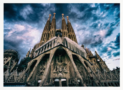 A dramatic, low-angle photograph of the Sagrada Familia in Barcelona, with its ornate stone facade and towering spires reaching up into a turbulent, dark blue and cloudy sky. The perspective emphasizes the grand scale and intricate detail of Gaudí's architecture. Print