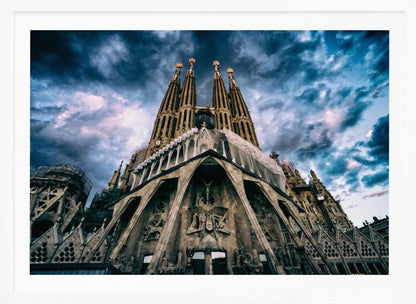 A dramatic, low-angle photograph of the Sagrada Familia in Barcelona, with its ornate stone facade and towering spires reaching up into a turbulent, dark blue and cloudy sky. The perspective emphasizes the grand scale and intricate detail of Gaudí's architecture. Print