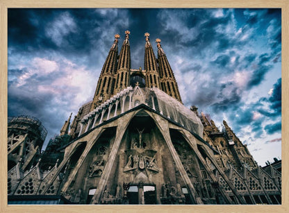 A dramatic, low-angle photograph of the Sagrada Familia in Barcelona, with its ornate stone facade and towering spires reaching up into a turbulent, dark blue and cloudy sky. The perspective emphasizes the grand scale and intricate detail of Gaudí's architecture. Print