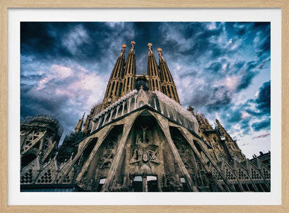 A dramatic, low-angle photograph of the Sagrada Familia in Barcelona, with its ornate stone facade and towering spires reaching up into a turbulent, dark blue and cloudy sky. The perspective emphasizes the grand scale and intricate detail of Gaudí's architecture. Print