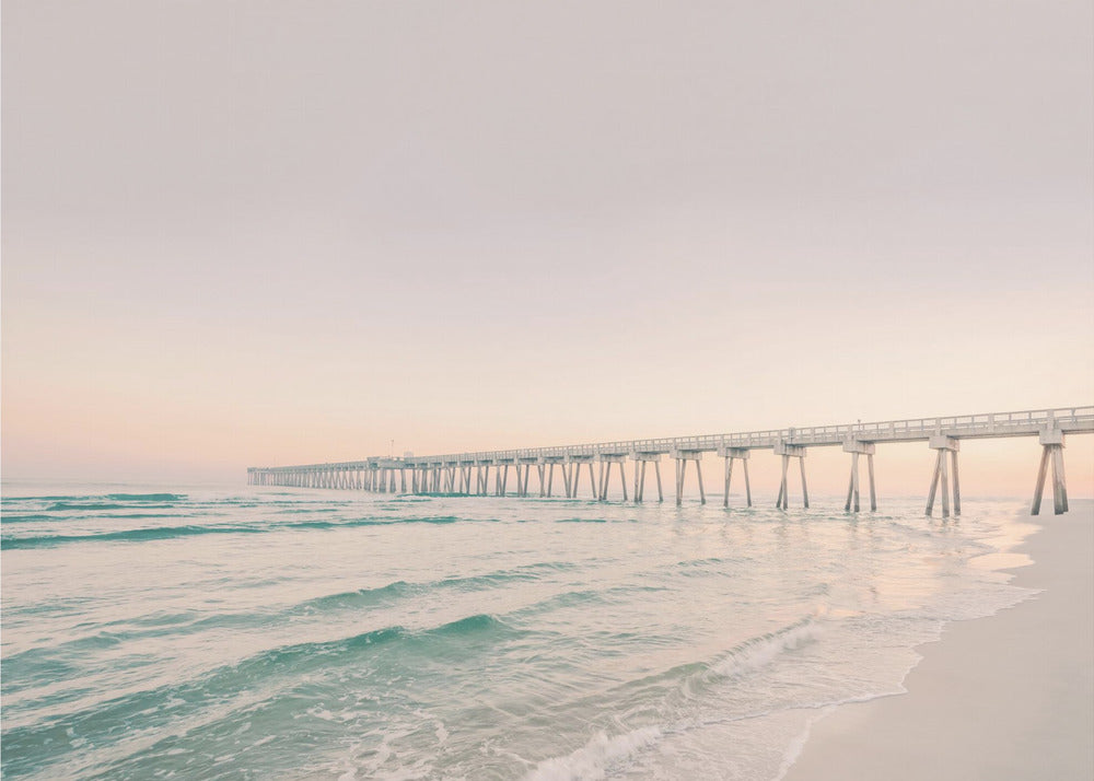 A tranquil beach scene with a long white pier extending into the turquoise ocean. The sky is a soft, pale pink, suggesting a serene sunrise or sunset, and gentle waves lap onto the sandy shore in the foreground. Poster