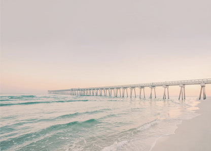 A tranquil beach scene with a long white pier extending into the turquoise ocean. The sky is a soft, pale pink, suggesting a serene sunrise or sunset, and gentle waves lap onto the sandy shore in the foreground. Poster