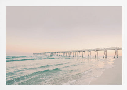 A tranquil beach scene with a long white pier extending into the turquoise ocean. The sky is a soft, pale pink, suggesting a serene sunrise or sunset, and gentle waves lap onto the sandy shore in the foreground. Poster