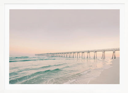 A tranquil beach scene with a long white pier extending into the turquoise ocean. The sky is a soft, pale pink, suggesting a serene sunrise or sunset, and gentle waves lap onto the sandy shore in the foreground. Poster