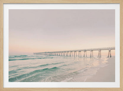 A tranquil beach scene with a long white pier extending into the turquoise ocean. The sky is a soft, pale pink, suggesting a serene sunrise or sunset, and gentle waves lap onto the sandy shore in the foreground. Poster