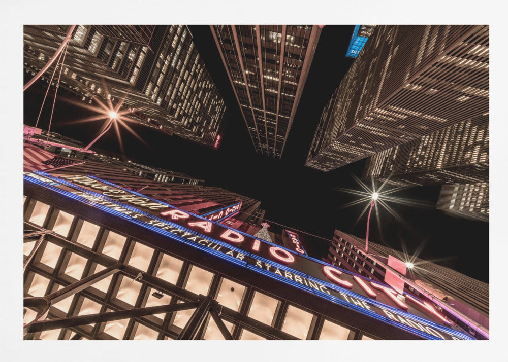A dramatic low-angle shot looking up at the iconic Radio City Music Hall marquee at night, surrounded by towering, illuminated skyscrapers against a dark sky. Decor