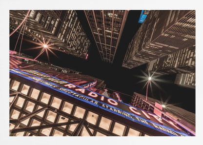 A dramatic low-angle shot looking up at the iconic Radio City Music Hall marquee at night, surrounded by towering, illuminated skyscrapers against a dark sky. Decor