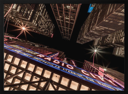 A dramatic low-angle shot looking up at the iconic Radio City Music Hall marquee at night, surrounded by towering, illuminated skyscrapers against a dark sky. Decor
