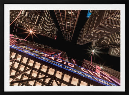 A dramatic low-angle shot looking up at the iconic Radio City Music Hall marquee at night, surrounded by towering, illuminated skyscrapers against a dark sky. Decor