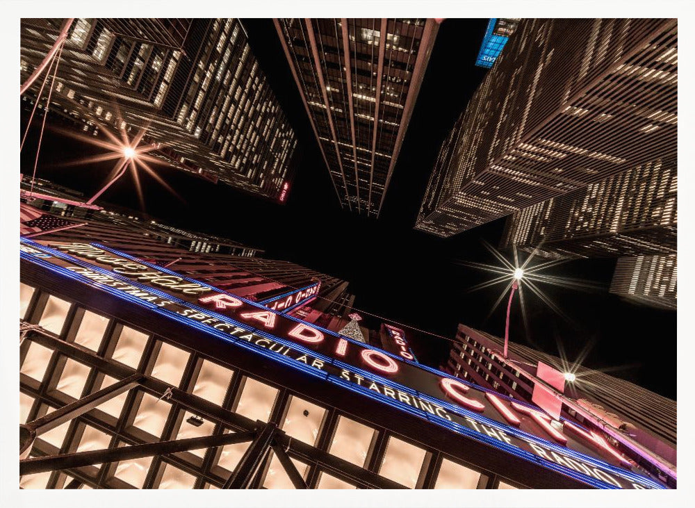 A dramatic low-angle shot looking up at the iconic Radio City Music Hall marquee at night, surrounded by towering, illuminated skyscrapers against a dark sky. Decor