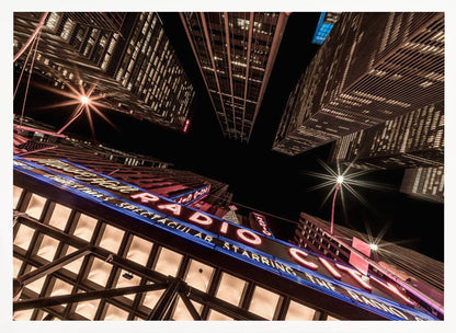 A dramatic low-angle shot looking up at the iconic Radio City Music Hall marquee at night, surrounded by towering, illuminated skyscrapers against a dark sky. Decor