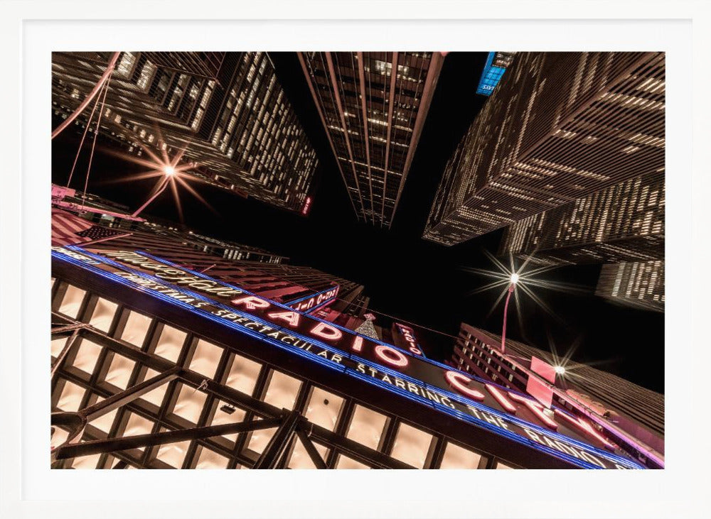 A dramatic low-angle shot looking up at the iconic Radio City Music Hall marquee at night, surrounded by towering, illuminated skyscrapers against a dark sky. Decor