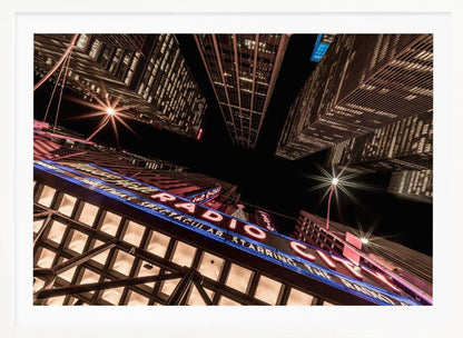 A dramatic low-angle shot looking up at the iconic Radio City Music Hall marquee at night, surrounded by towering, illuminated skyscrapers against a dark sky. Decor