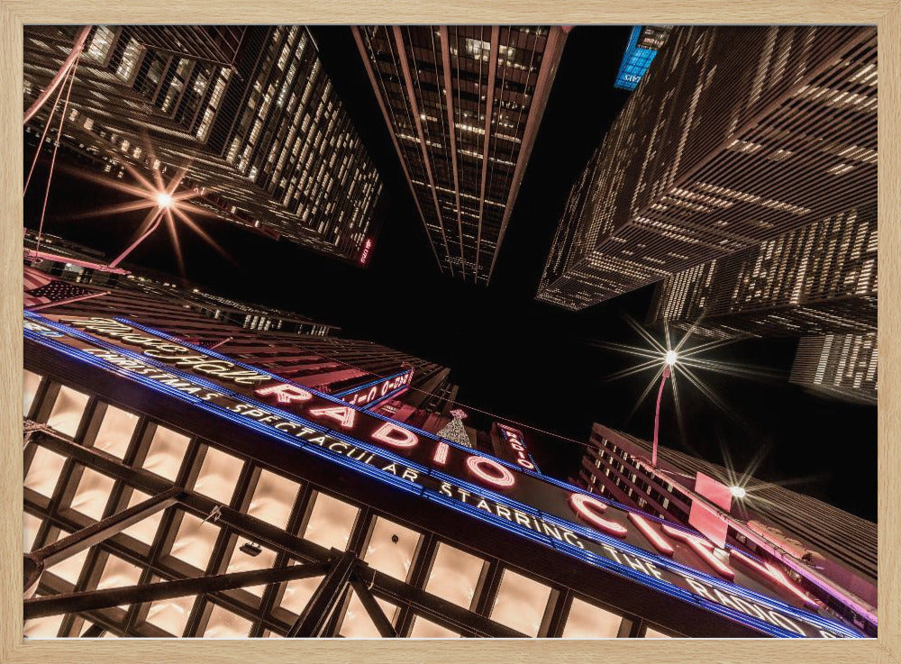 A dramatic low-angle shot looking up at the iconic Radio City Music Hall marquee at night, surrounded by towering, illuminated skyscrapers against a dark sky. Decor