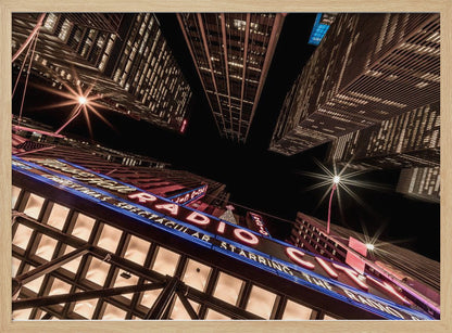 A dramatic low-angle shot looking up at the iconic Radio City Music Hall marquee at night, surrounded by towering, illuminated skyscrapers against a dark sky. Decor
