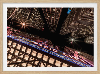 A dramatic low-angle shot looking up at the iconic Radio City Music Hall marquee at night, surrounded by towering, illuminated skyscrapers against a dark sky. Decor