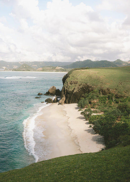 A high-angle photograph of a secluded tropical beach, with turquoise waves gently washing onto the white sand. A lush green cliff covered in grass and small trees frames the cove, with distant rolling hills visible under a soft, cloudy sky. Decor