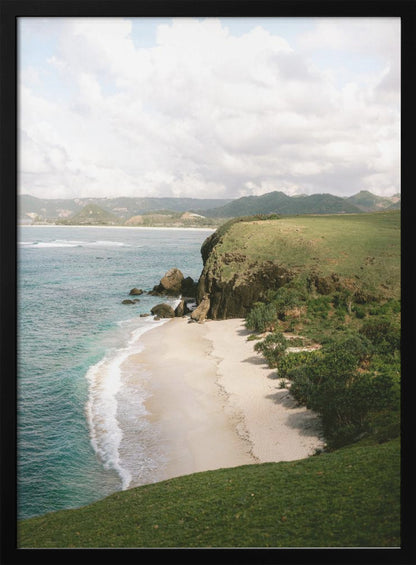A high-angle photograph of a secluded tropical beach, with turquoise waves gently washing onto the white sand. A lush green cliff covered in grass and small trees frames the cove, with distant rolling hills visible under a soft, cloudy sky. Decor