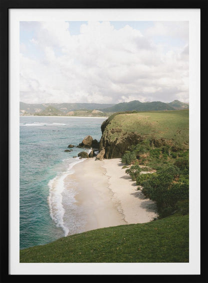 A high-angle photograph of a secluded tropical beach, with turquoise waves gently washing onto the white sand. A lush green cliff covered in grass and small trees frames the cove, with distant rolling hills visible under a soft, cloudy sky. Decor