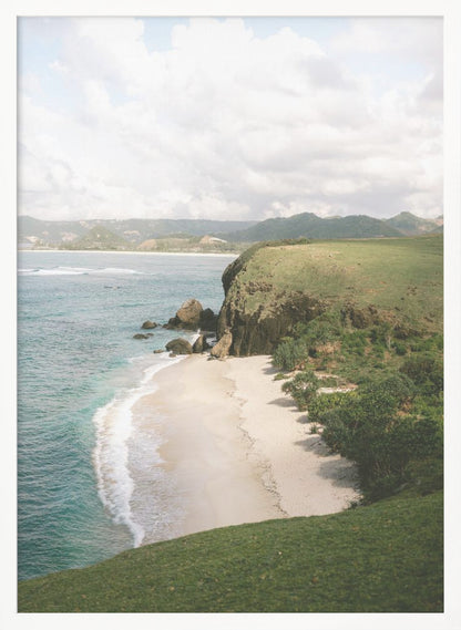 A high-angle photograph of a secluded tropical beach, with turquoise waves gently washing onto the white sand. A lush green cliff covered in grass and small trees frames the cove, with distant rolling hills visible under a soft, cloudy sky. Decor