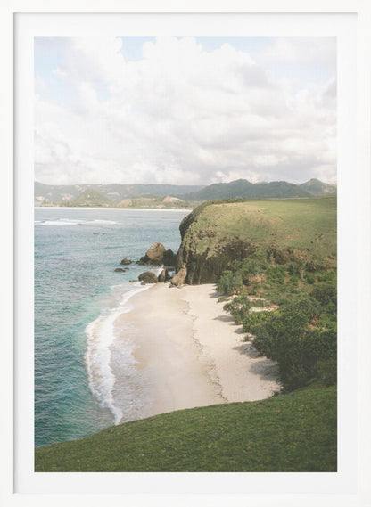 A high-angle photograph of a secluded tropical beach, with turquoise waves gently washing onto the white sand. A lush green cliff covered in grass and small trees frames the cove, with distant rolling hills visible under a soft, cloudy sky. Decor
