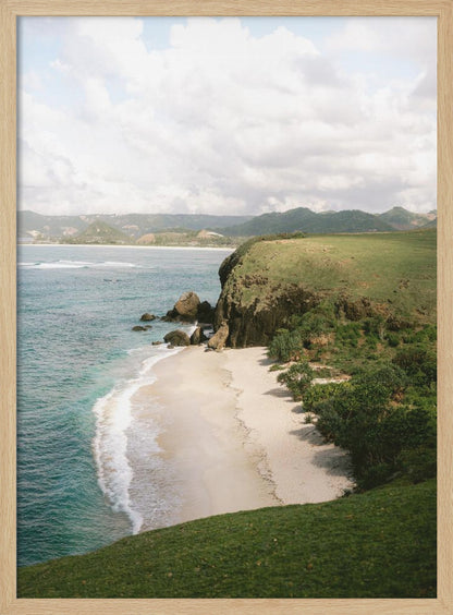 A high-angle photograph of a secluded tropical beach, with turquoise waves gently washing onto the white sand. A lush green cliff covered in grass and small trees frames the cove, with distant rolling hills visible under a soft, cloudy sky. Decor