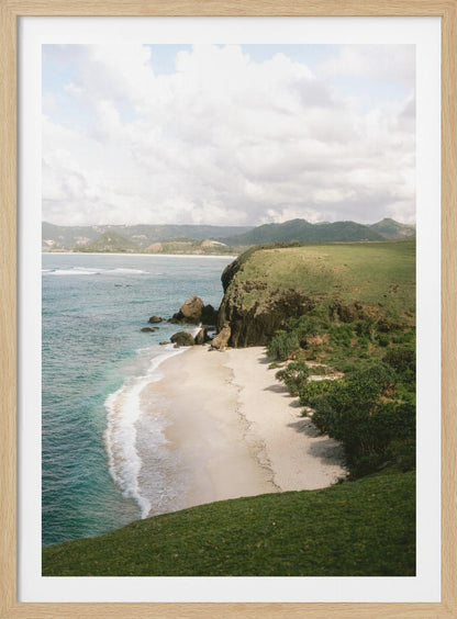 A high-angle photograph of a secluded tropical beach, with turquoise waves gently washing onto the white sand. A lush green cliff covered in grass and small trees frames the cove, with distant rolling hills visible under a soft, cloudy sky. Decor