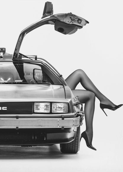 A black and white studio photograph of a DeLorean car with its gull-wing door open, featuring a woman's legs in black stockings and stilettos elegantly emerging from the passenger side. Decor