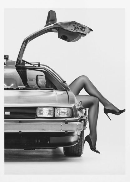 A black and white studio photograph of a DeLorean car with its gull-wing door open, featuring a woman's legs in black stockings and stilettos elegantly emerging from the passenger side. Decor